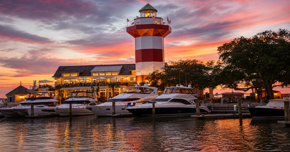 Harbour Town Lighthouse at sunset with the new Quarterdeck restaurant, yachts in the marina, and golden sky over Calibogue Sound on Hilton Head Island